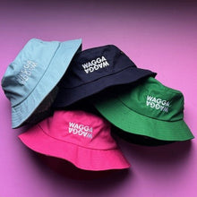 Load image into Gallery viewer, Four bucket hats in green, pink, navy, and light blue, arranged in a circle on a vibrant pink background. Each hat features the brand name 'WAGGA WAGGA' embroidered in white on the front.
