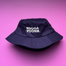 Load image into Gallery viewer, A dark navy blue bucket hat with 'WAGGA' embroidered in white on the front, displayed on a solid pink background.
