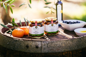 Two jars of olives, two mandarins, a bottle of olive oil and olives in a bowl styled on top of a wine barrel. 